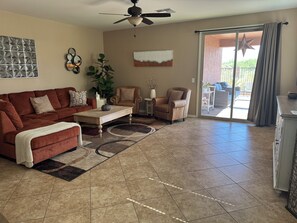 A living room with a brown sofa, a coffee table, and a chair. There is a ceiling fan, a large window with a view, and a sliding glass door leading to an outdoor area. - Dove Mountain Perserve, gated, dog friendly home. (Marana)