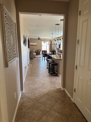 A hallway leading to a dining area with a ceiling fan, a kitchen with bar stools, and a door on the right.