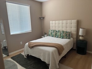 A bedroom with a bed, a bedside lamp, a floor lamp, and a window with blinds.