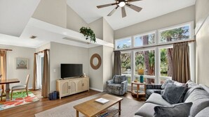 A spacious living room with a ceiling fan, a television on a wooden cabinet, a sofa, and large windows with curtains.