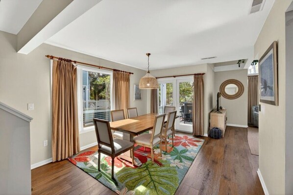 A dining area with a wooden table and chairs, a large window with curtains, a hanging light fixture, and a colorful rug.