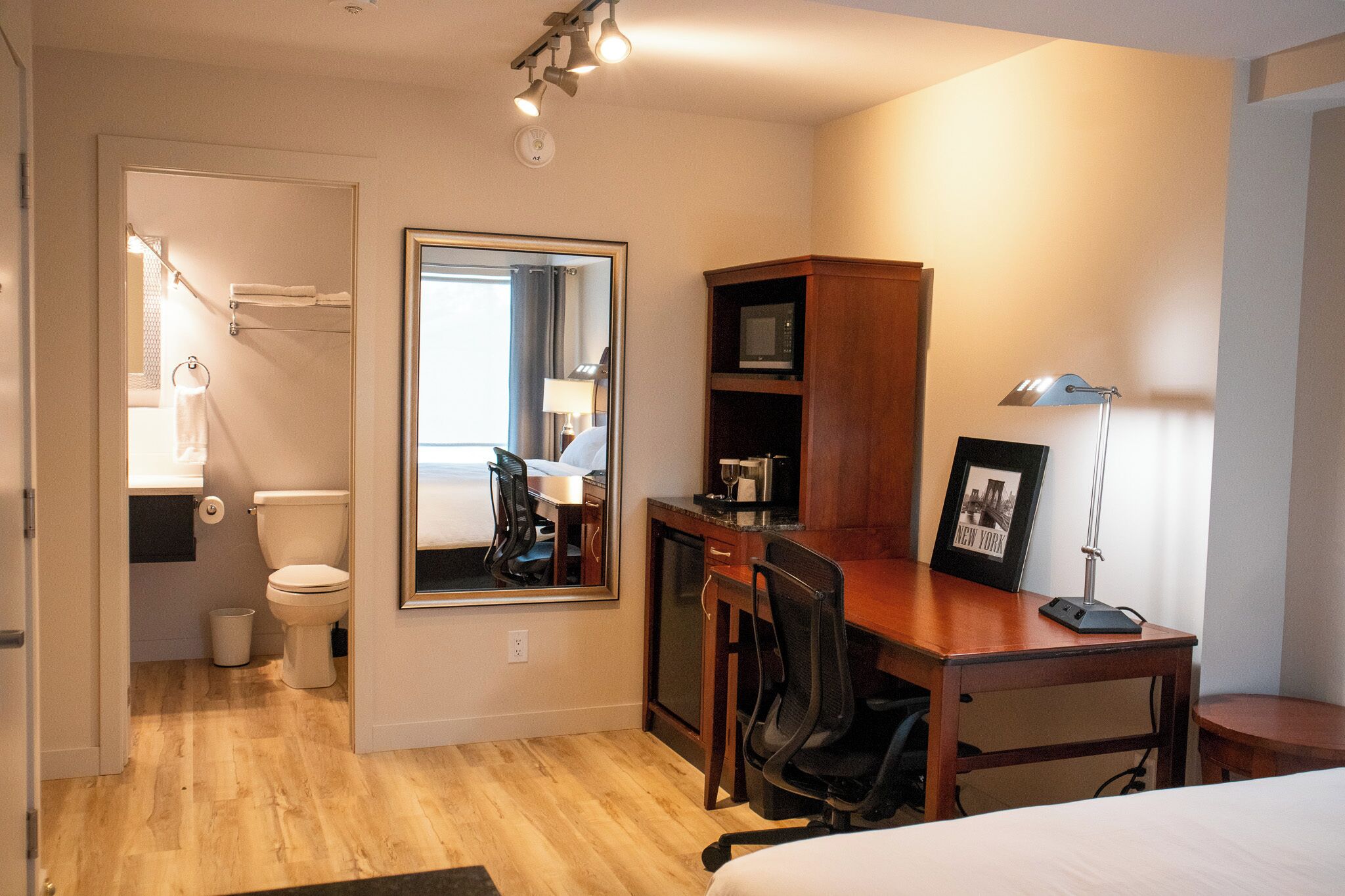 A hotel room with a wooden desk, a chair, a bed, a desk lamp, a framed picture, a microwave, and a large mirror.