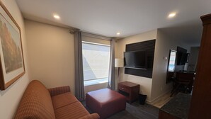 A hotel room with a sofa, a small table, a TV mounted on the wall, a window with curtains, and a view of the outside. - Auberge Le Chardo Inn (Alfred)