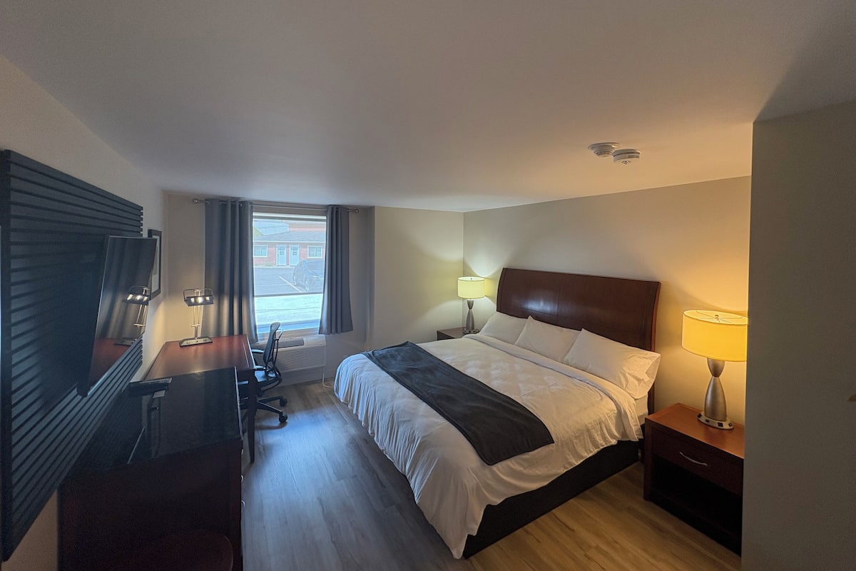 A hotel room with a bed, bedside tables, a desk, a chair, and a large window.