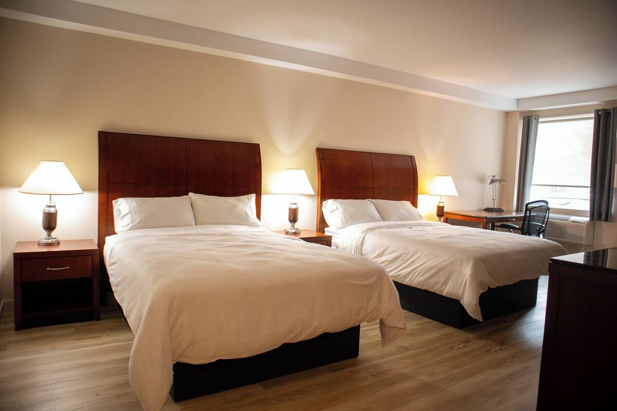 A hotel room with two beds, wooden headboards, bedside tables with lamps, a desk, and a chair.