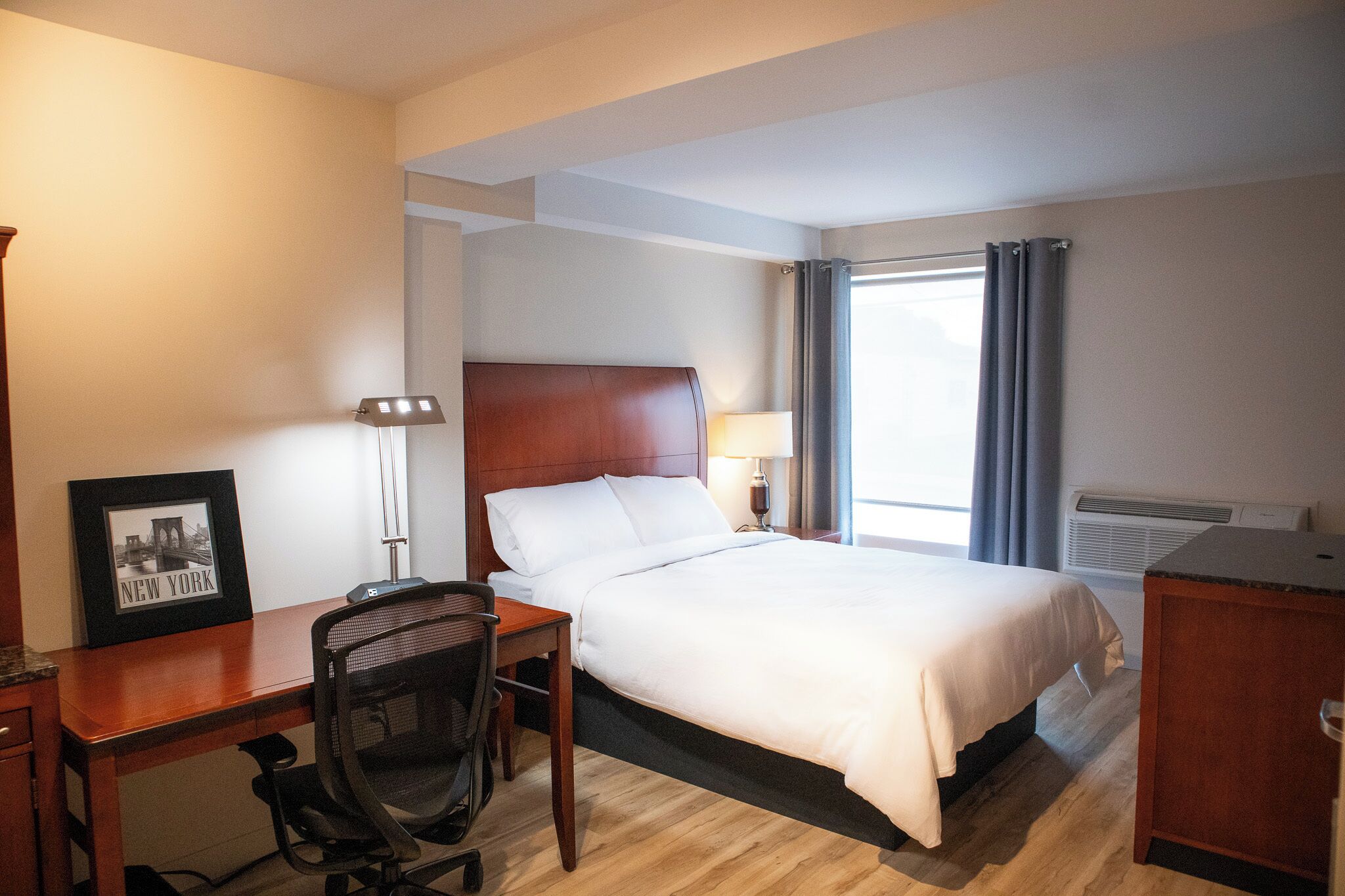 A hotel room with a bed, desk, chair, lamp, and a framed picture on the wall.