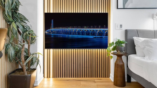 A modern hotel room with a large flat-screen TV mounted on a wooden slat wall, a grey sofa, a small wooden side table, and a potted plant.