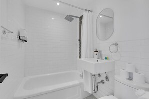 Ein modernes Badezimmer mit einer weißen Badewanne, einer Dusche mit Glastür, einem Waschbecken mit Spiegel und Toilettenpapier an der Wand.