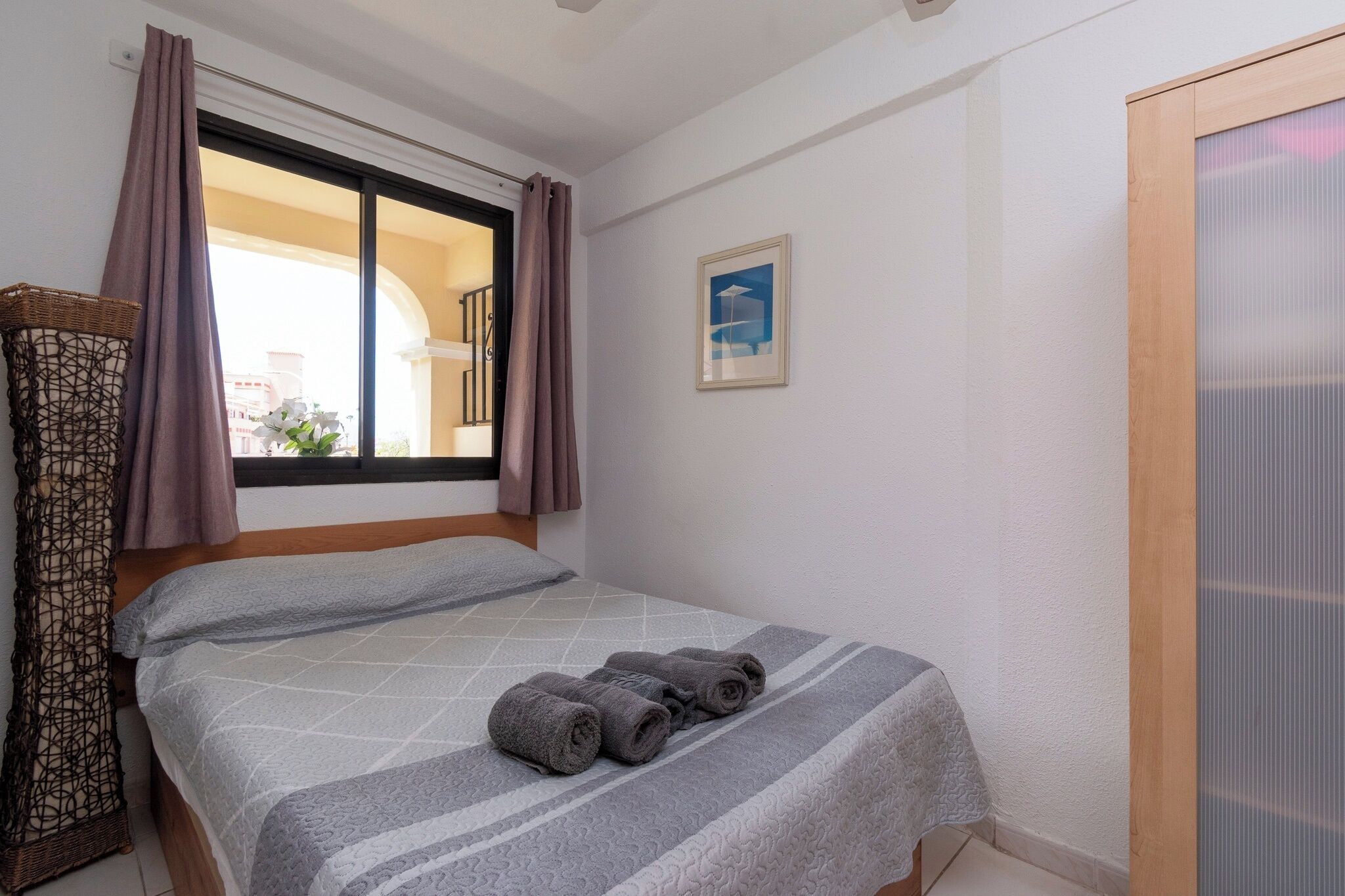 Een slaapkamer met een bed, een raam met gordijnen, een lijst met een foto aan de muur en een houten deur.