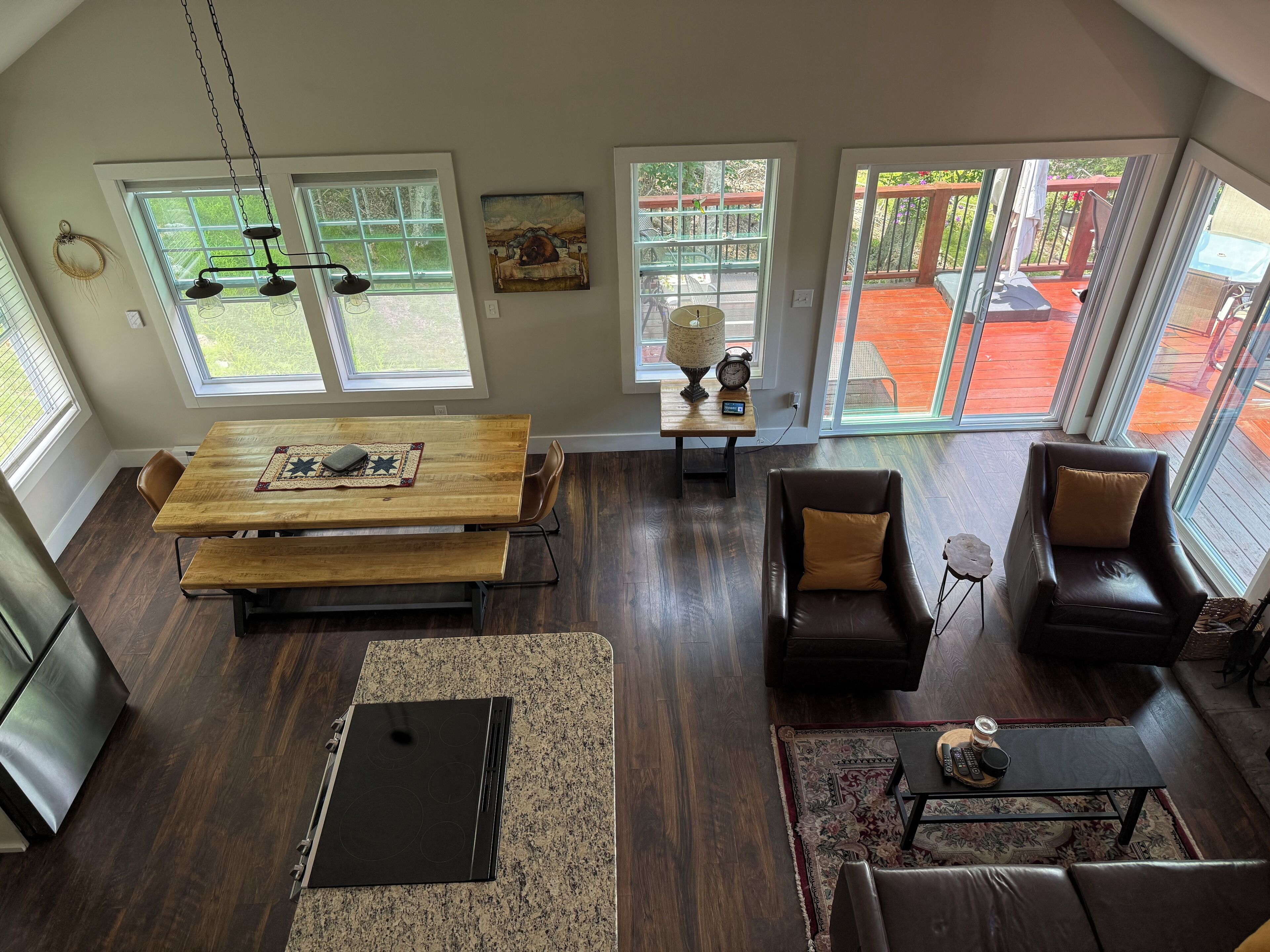A spacious living area with a wooden dining table, a comfortable sofa, and a kitchen with a built-in stove. Large windows offer views of a backyard.