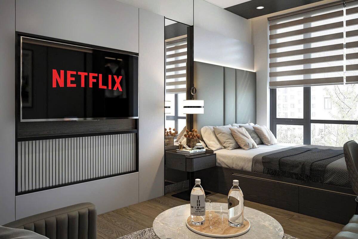 Une chambre moderne dotée d’une grande télévision affichant le logo « NETFLIX », d’un lit avec une tête de lit, de tables de chevet et d’une table basse ronde sur laquelle sont posées des bouteilles d’eau.