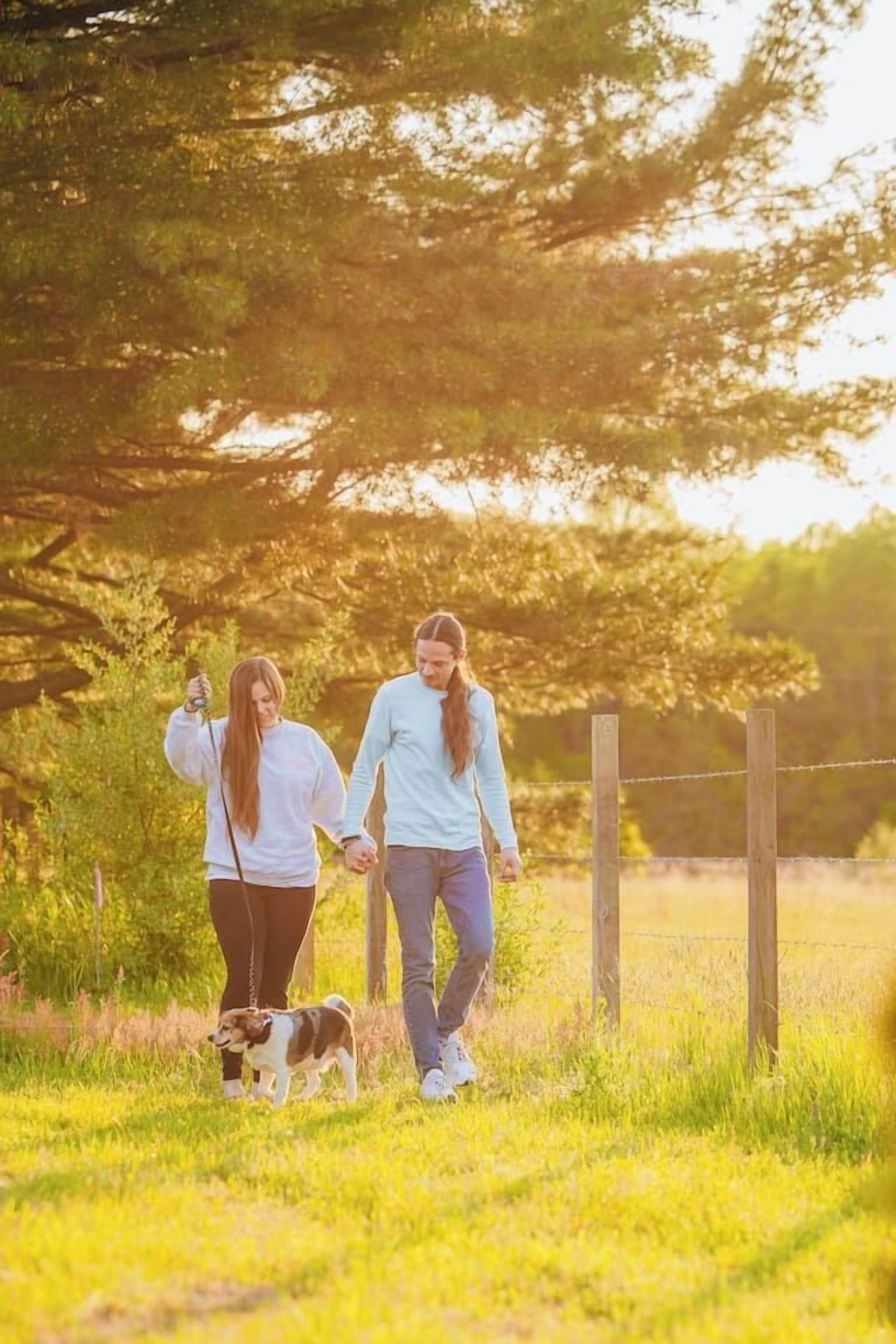 Due persone passeggiano con un cane in una zona erbosa all'aperto, con alberi e una recinzione.
