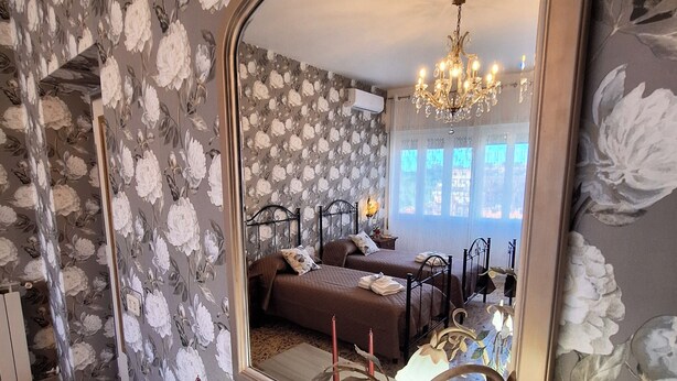 A room with floral wallpaper, a large mirror reflecting a bed and a chandelier, and a decorative plant in the foreground. - A Casa di Noele (Roma)