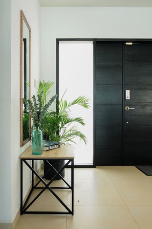 Una entrada moderna con una puerta negra, una consola de madera con un florero de plantas verdes, un espejo y un piso de baldosas.