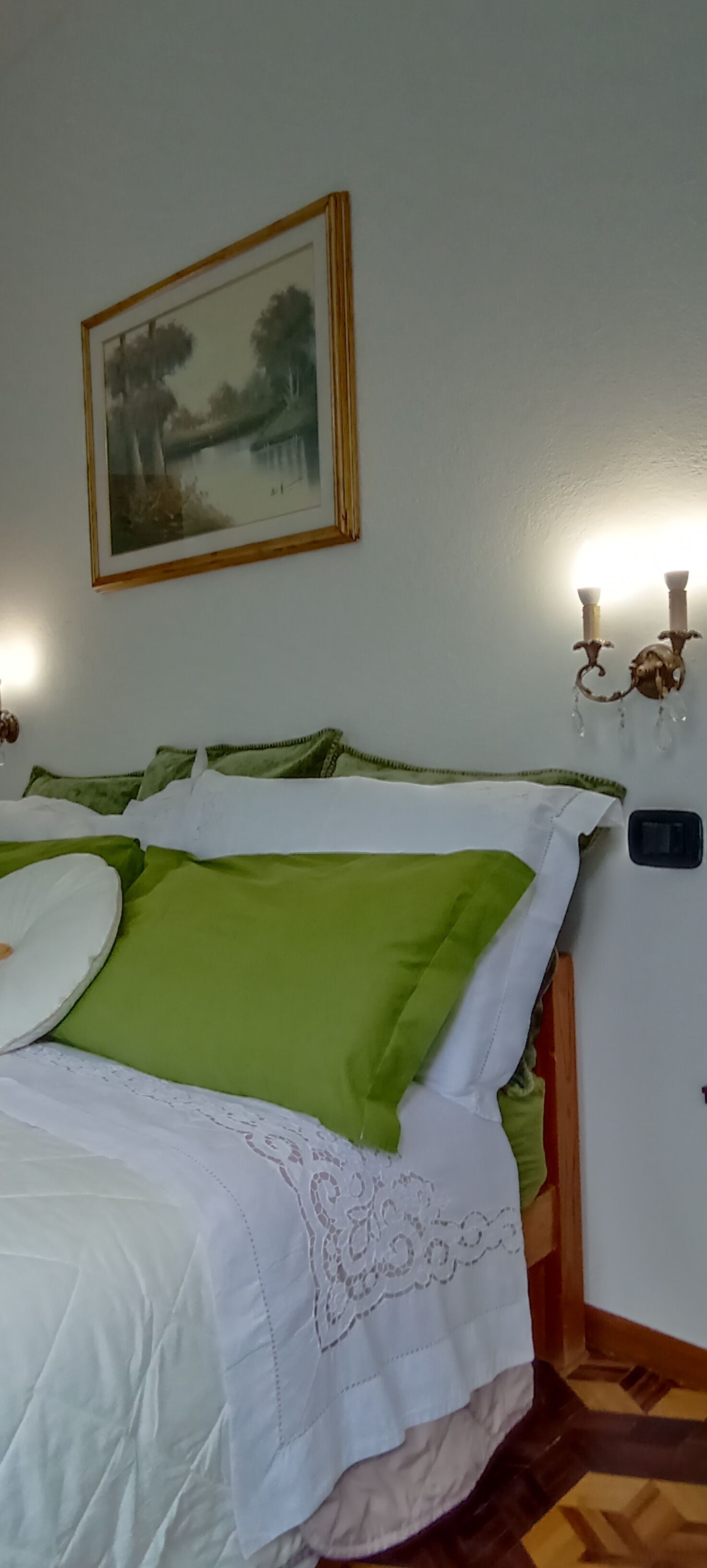 Una cama bien hecha con ropa de cama verde y blanca, un cuadro de paisaje en la pared y una lámpara de pared.