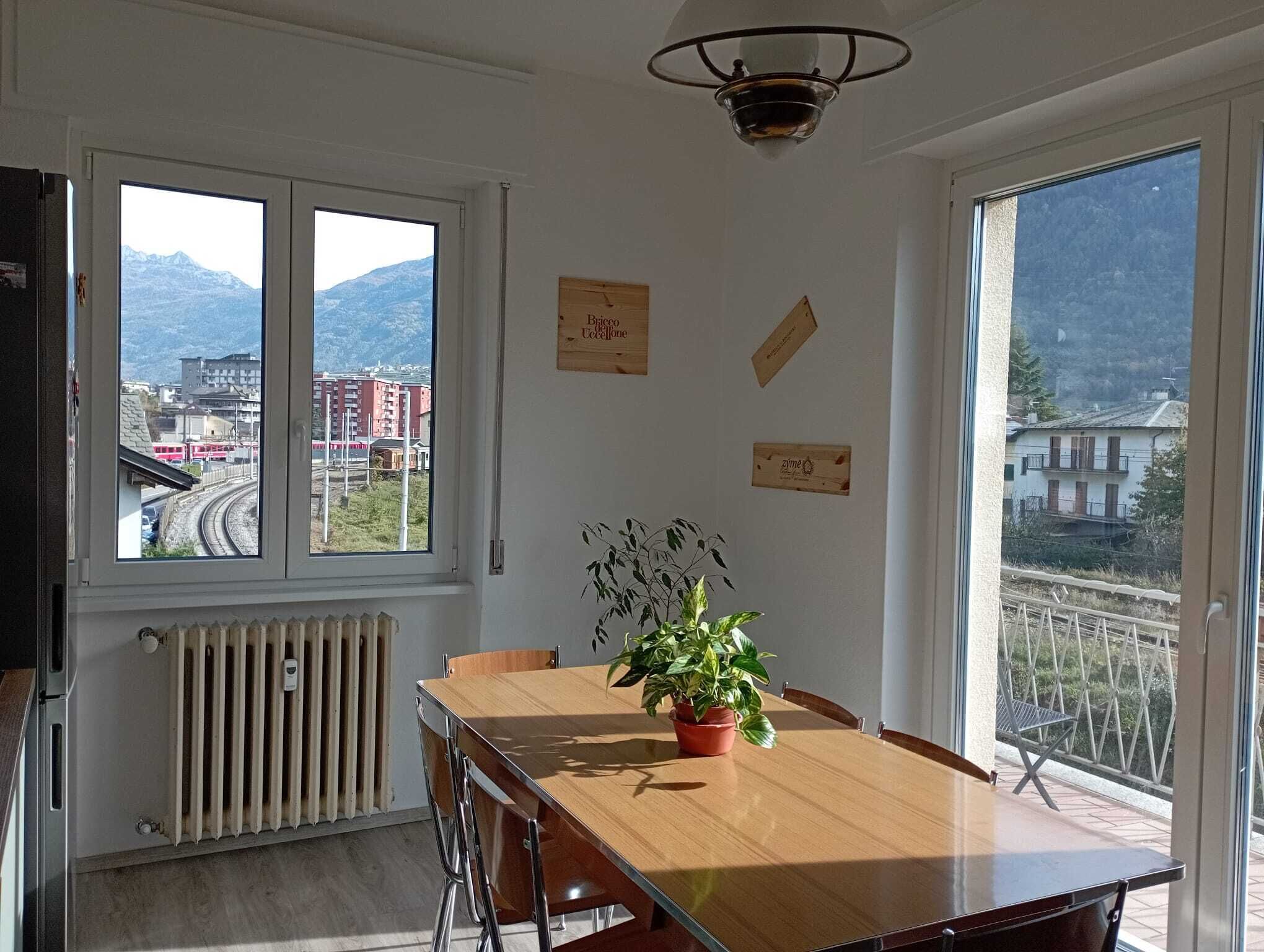Zona pranzo con tavolo in legno, sedie, una pianta in vaso e vista sulle montagne e sugli edifici attraverso ampie finestre.