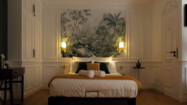 Une chambre à coucher avec un grand lit, une table de chevet, un tableau représentant une végétation tropicale et deux lampes fixées au mur.