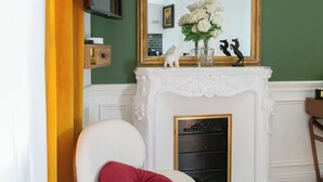 Une pièce avec une chaise blanche, un coussin rouge, une cheminée avec un manteau de couleur blanche et un grand miroir décoratif.
