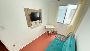 Una habitación con un sofá color turquesa, un televisor montado, un escritorio pequeño blanco y una ventana con persianas.
