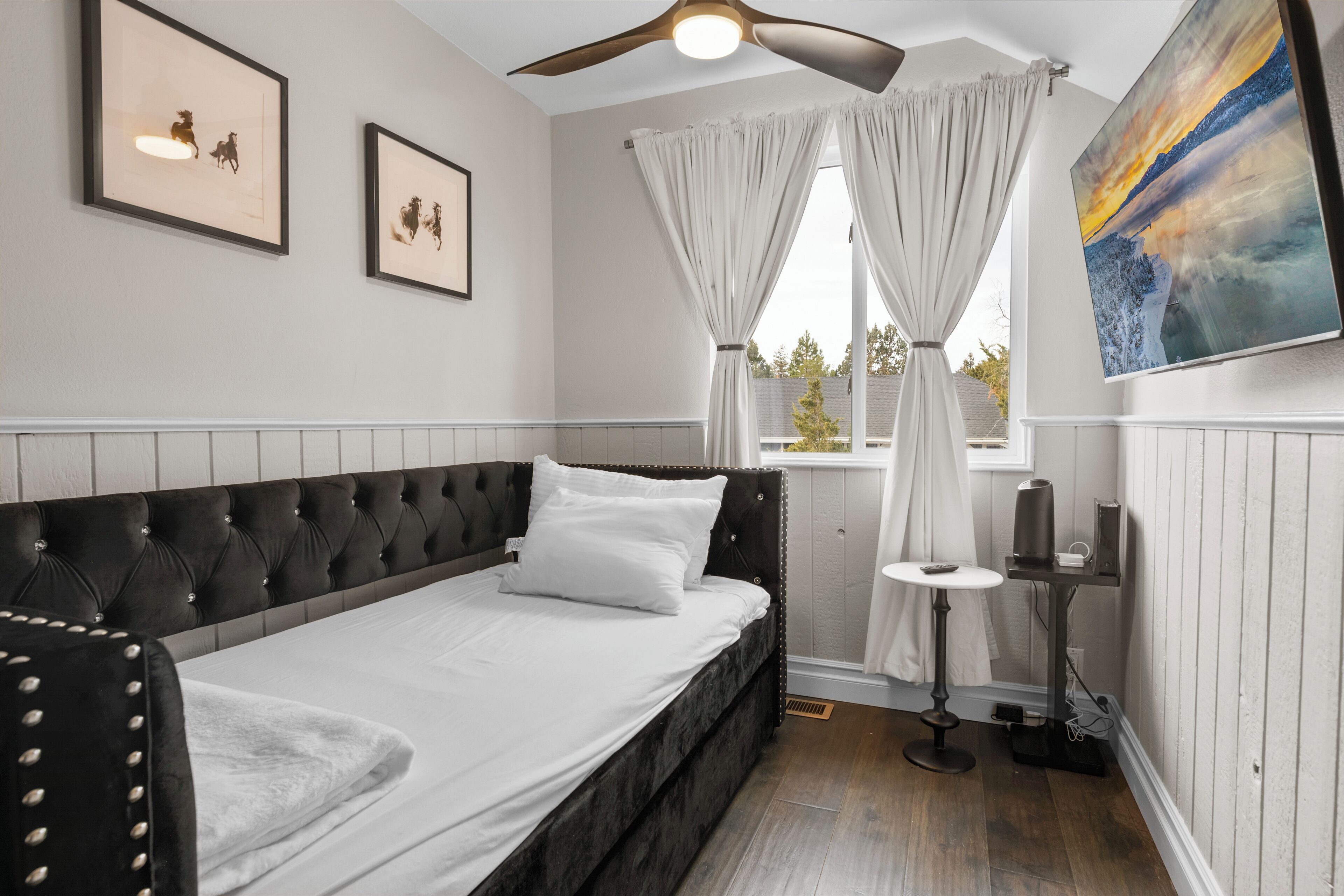 A bedroom with a bed, ceiling fan, artwork, and a window with curtains.