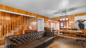 A cozy living room with a wooden staircase, a leather sofa, a dining area with a long table, and a kitchen in the background. - Bunny's Ranch (Big Bear City)