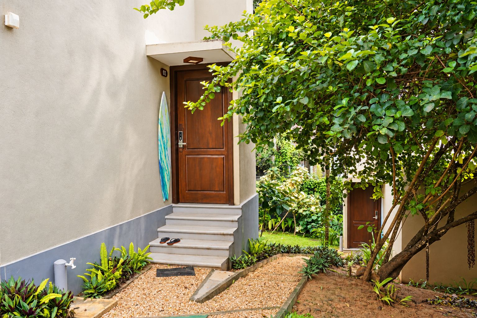 A residential building with a wooden door, a small set of steps leading to the entrance, and a garden area with various plants.