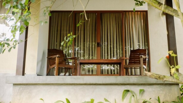 Un balcón con dos sillas de madera y una mesa, cortinas, y vista al interior a través de una ventana.