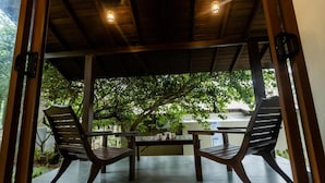 Un espacio al aire libre con dos sillas de madera y una mesa, cubierto por un techo de madera con luces colgantes, rodeado de follaje.