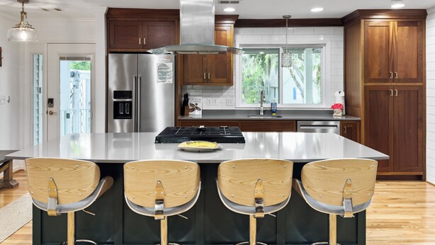 Uma cozinha moderna com ilha central, banquetas de madeira, eletrodomésticos de aço inoxidável e uma grande janela que oferece luz natural.