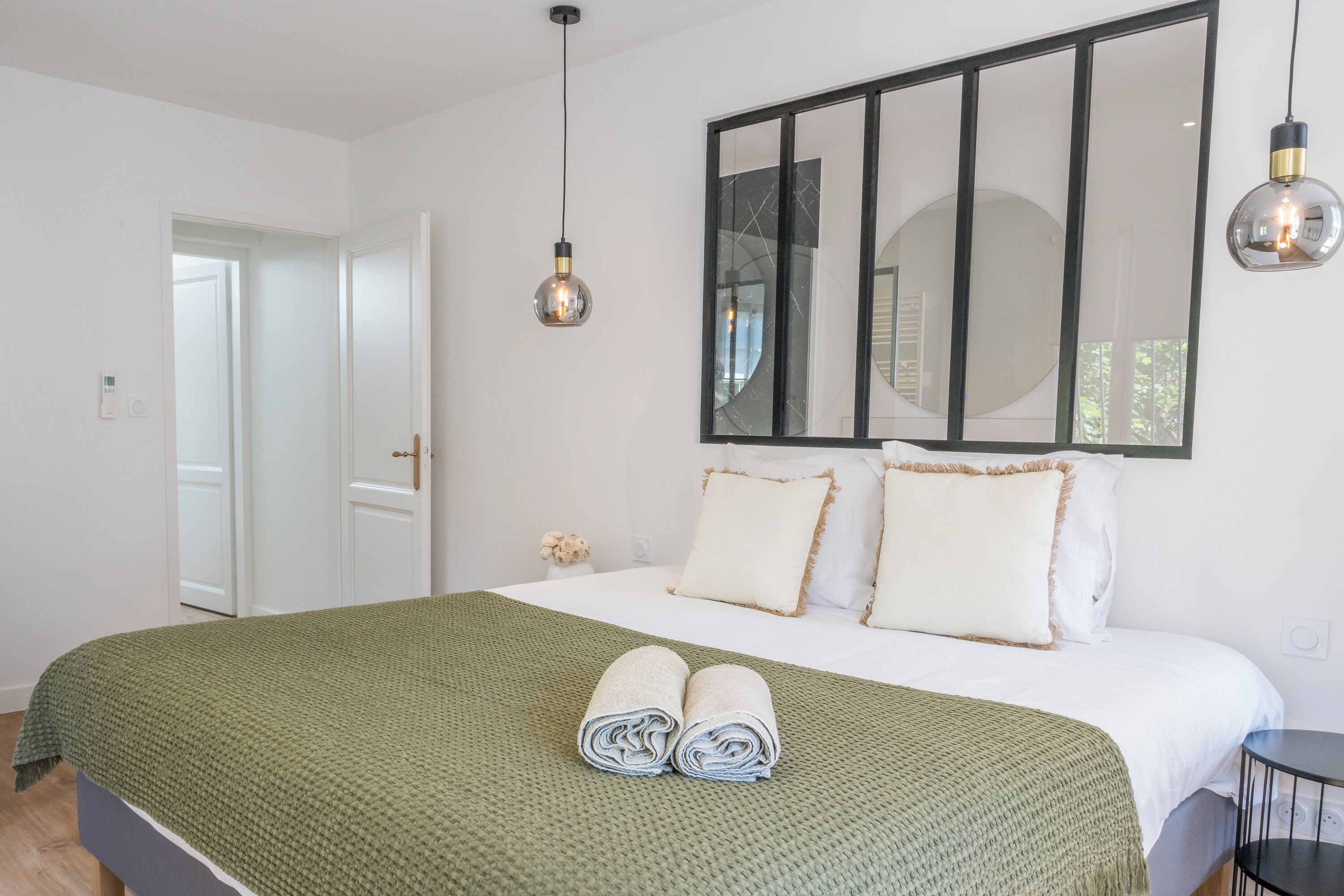 Een net opgemaakt bed met een groene deken, twee kussens en opgerolde handdoeken op een witte sprei. Daarboven hangt een grote spiegel en twee hangende hanglampen.