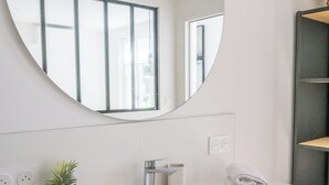 A modern bathroom with a white sink, a round mirror, and a potted plant. - Les Tamaris (SAINTE TERRE)