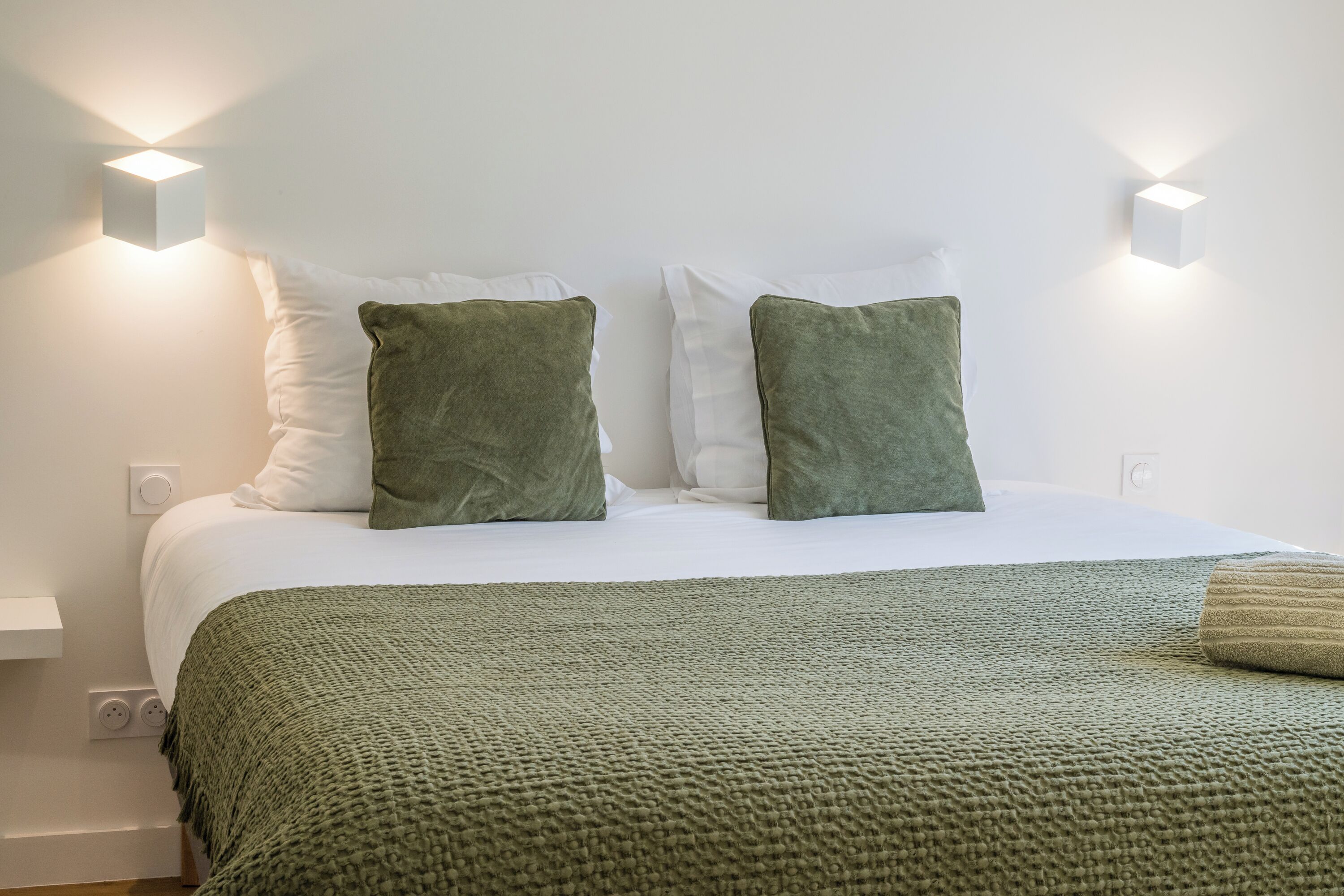A neatly made bed with a green blanket and two green pillows, a white wall, and two wall-mounted light fixtures.