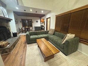 A living room with a sectional sofa, a wooden coffee table, a fireplace, and a kitchen area in the background.