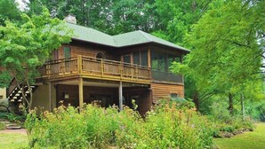 A two-story log cabin with a balcony, surrounded by lush greenery and a stone retaining wall. - Tranquil Cardinal Hill Cabin- Perfect for Relaxation & Exploration. Pet Friendly (Sperryville)