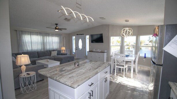 A modern kitchen with a central island, a dining area with a white table and chairs, a grey sofa set, and a ceiling fan. - Pond View 2BR 2BA Beach Escape (Fort Myers Beach)