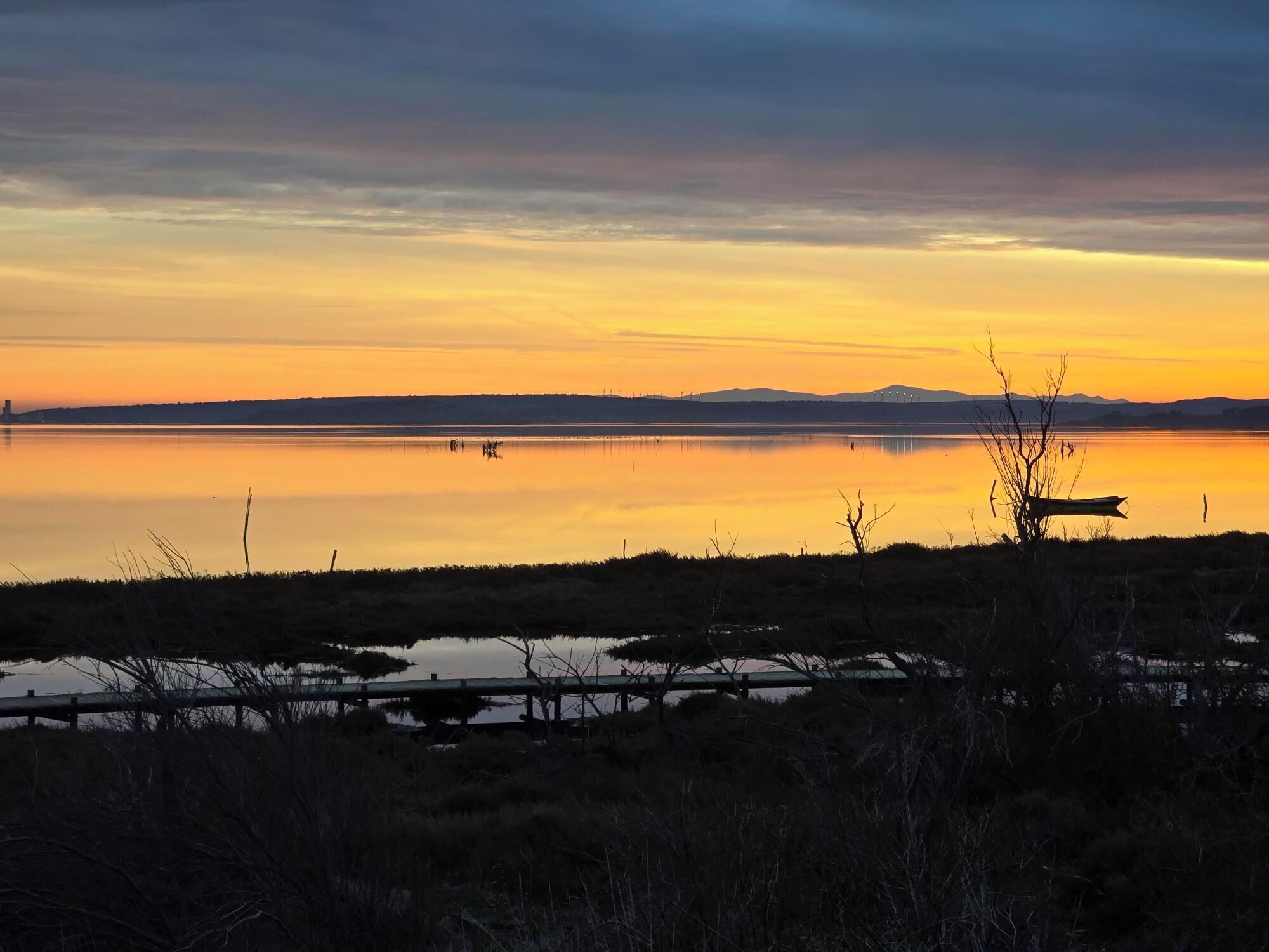Een rustig meer bij zonsondergang met een houten steiger en schaarse begroeiing op de voorgrond.