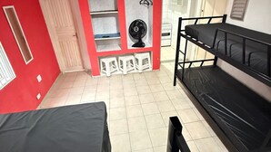 Un dormitorio con literas, ventilador de techo, ventilador pequeño y ventana con persianas.