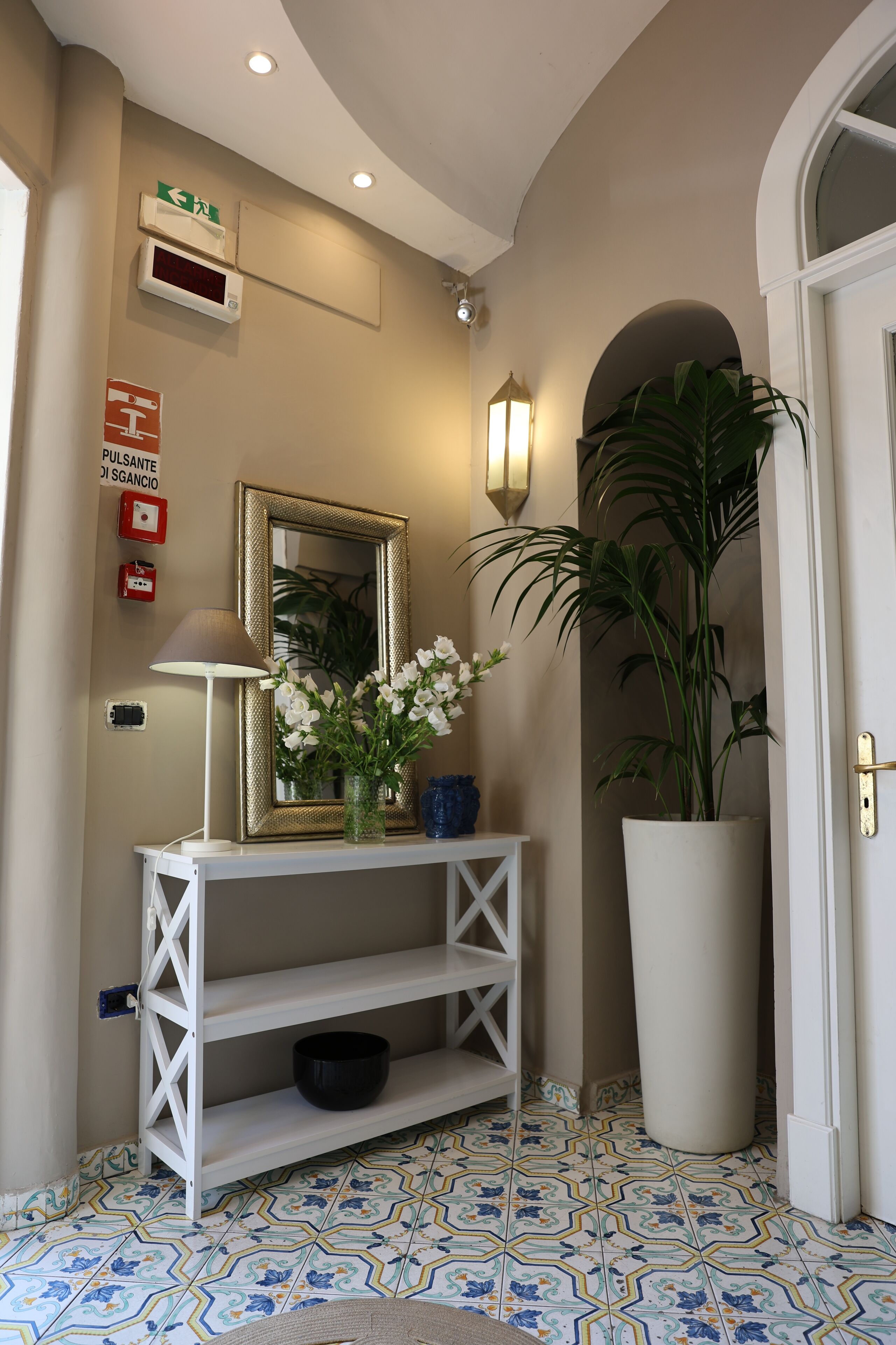 Un couloir avec une console blanche, un miroir, une plante en pot et un sol carrelé.