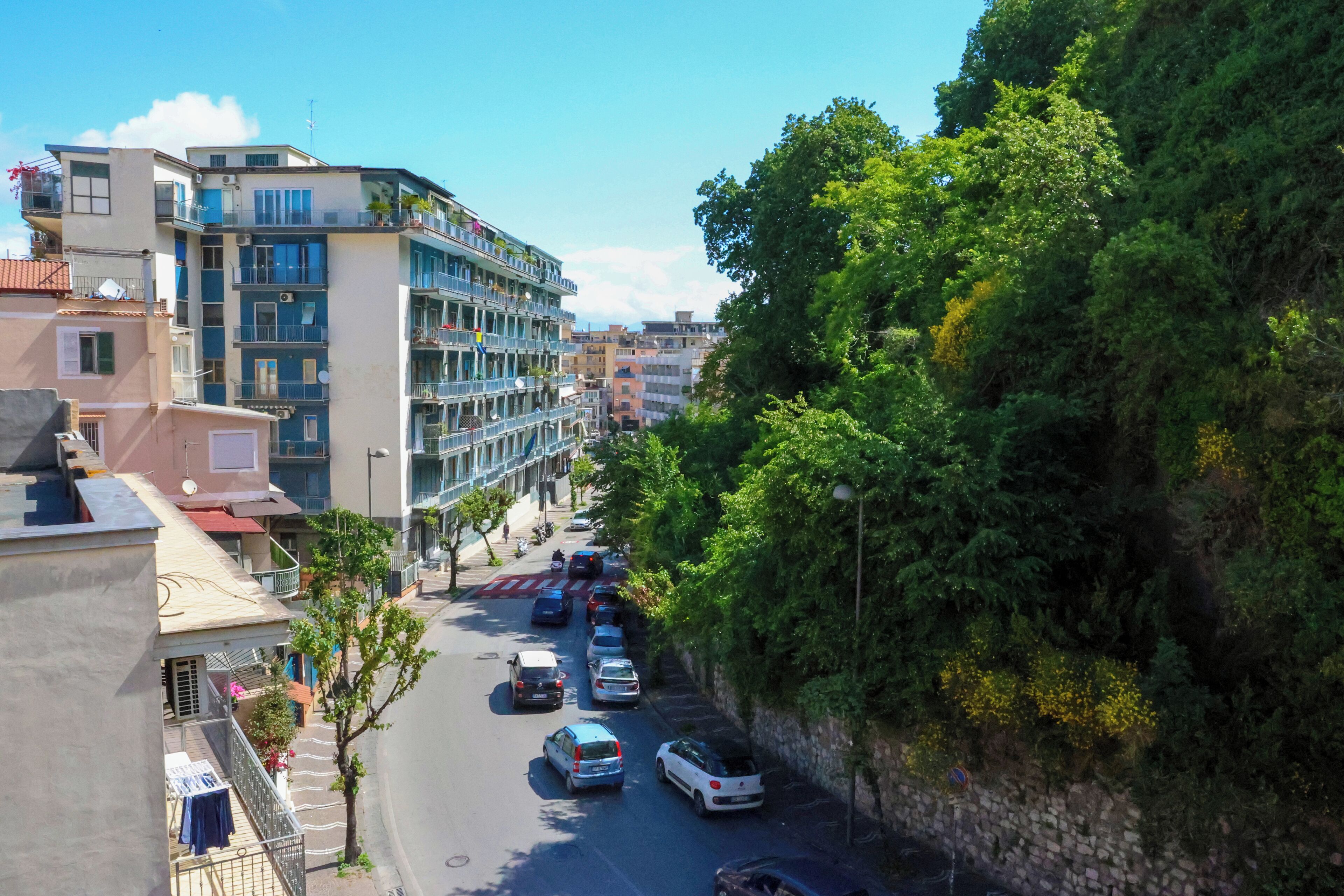 A multi-story building with balconies, a street with parked cars, and a tree-lined hillside.