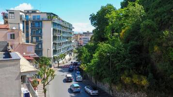 A multi-story building with balconies, a street with parked cars, and a tree-lined hillside.