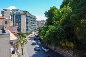 Un edificio de varios pisos con balcones, una calle con coches aparcados y una ladera arbolada.