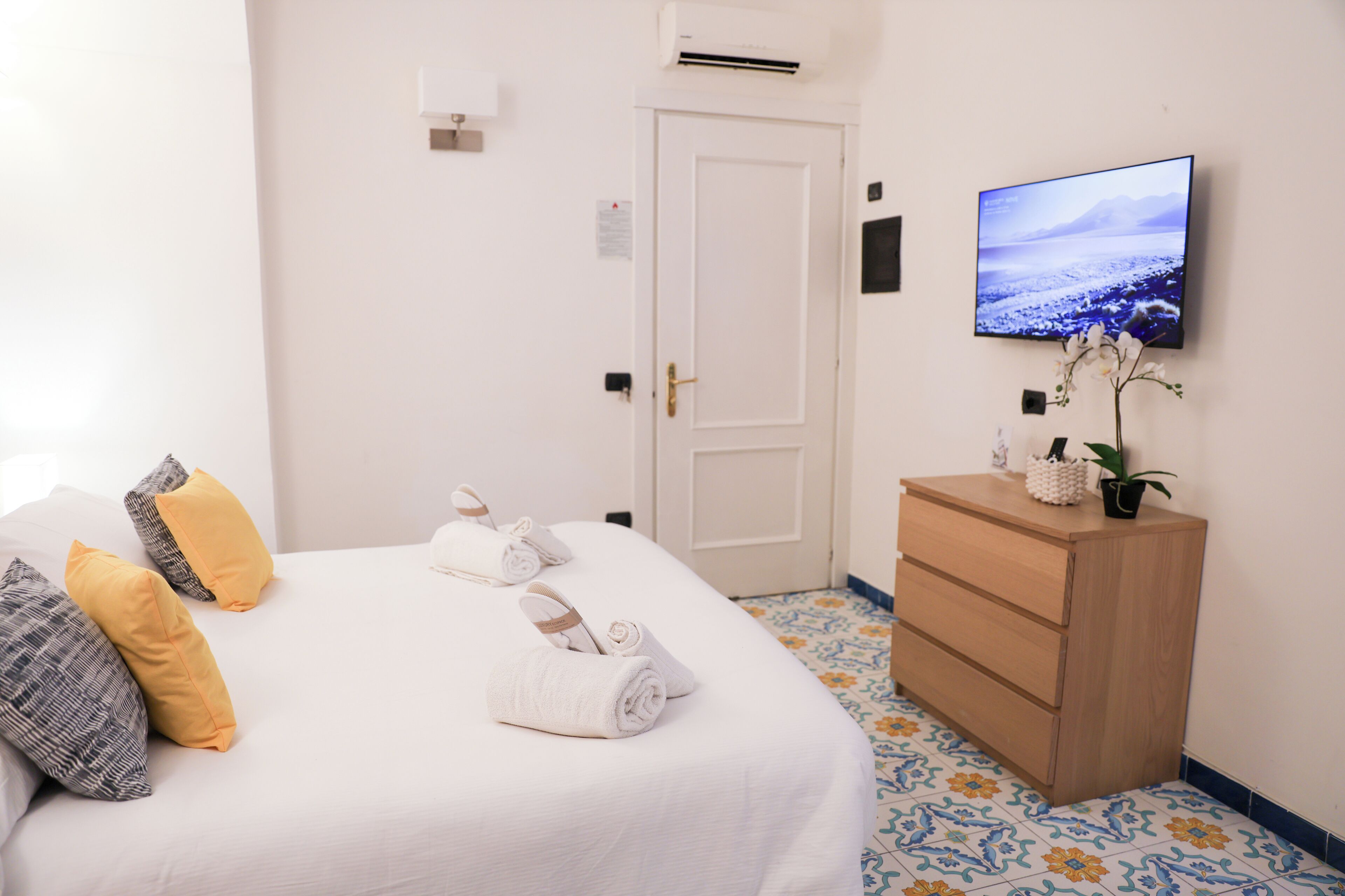 Una cama bien hecha con almohadas blancas y amarillas, un aparador de madera con una planta en maceta y un televisor de pantalla plana en la pared.