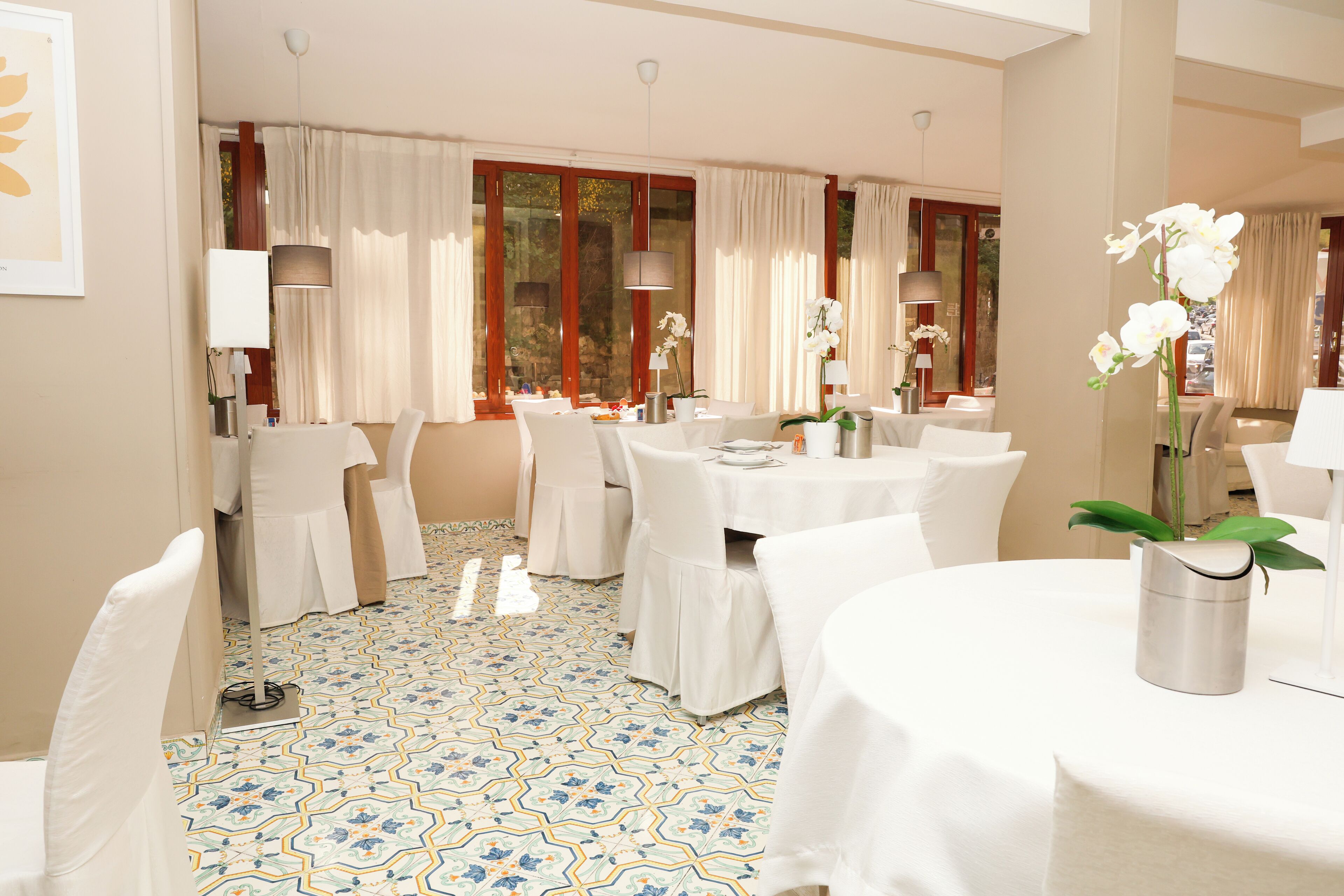 Uma sala de jantar de hotel com mesas redondas, cadeiras brancas e piso estampado.