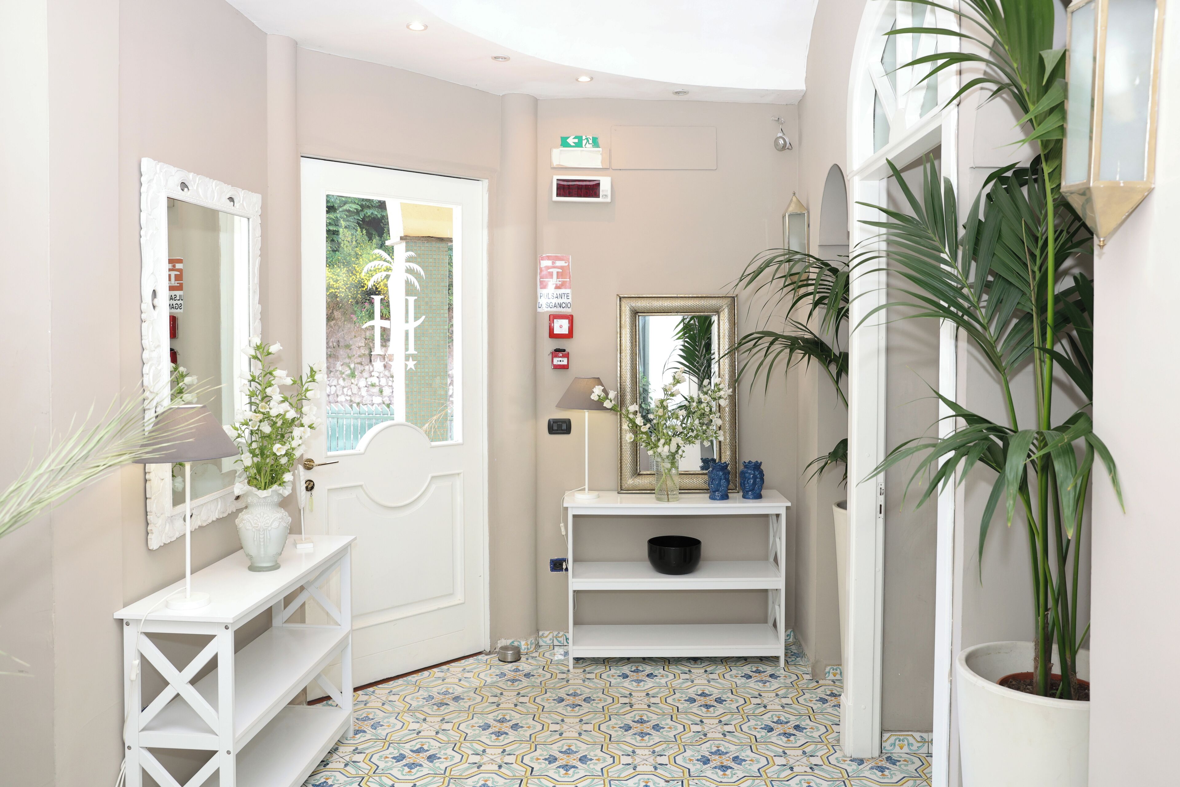 Um corredor com porta branca, mesa consola branca com um vaso de flores, um espelho e uma estante com objetos decorativos.