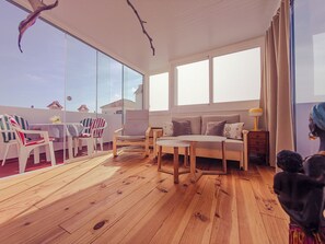 A spacious room with a wooden floor, a sofa, a table, and a chair. There are large windows and a view of the sky. - Marino Home: 2-bedroom Townhome w Patio & Swimming pool (Torrevieja)