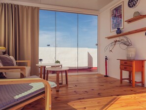 Une pièce comprenant un lit, une petite table, une chaise, une table d’appoint en bois et une grande porte-fenêtre coulissante donnant sur un espace extérieur.