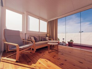 A balcony with a wooden floor, a sofa, a chair, a small table, and potted plants.