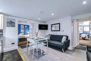 A modern living room with a sofa, a dining table with chairs, a flat-screen TV, and a cityscape wall art.