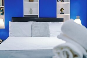 A neatly made bed with white linens and a textured pillow, a bedside lamp, and a bookshelf with various items including books and decorative objects.