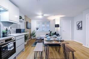 A modern kitchen with a dining area, featuring built-in appliances, a dining table set for four, and a window with blinds.